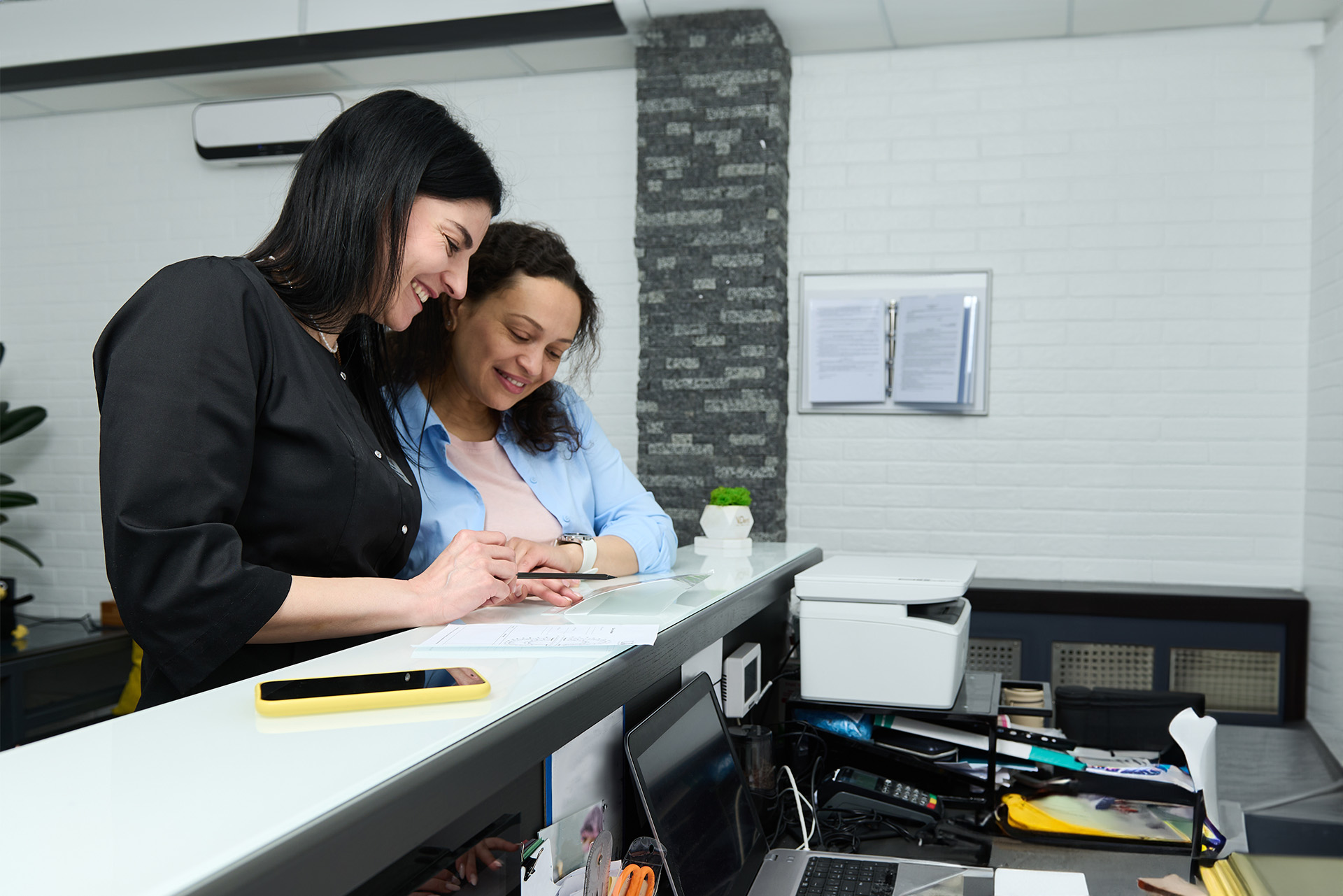 The image shows two individuals, presumably in a professional setting such as an office or a service establishment, with one person seated at a desk and the other standing nearby.