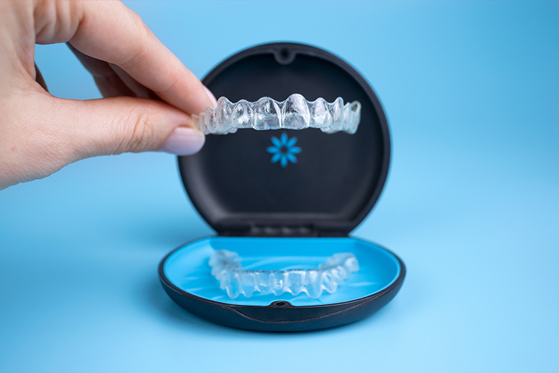 An image of a person s hand holding a transparent plastic dental retainer with a blue lid, showcasing the clear, tooth-shaped structure inside.