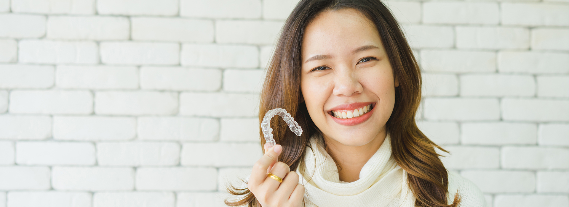 The image shows a smiling woman with a ring on her finger, holding up a small object towards the camera. She is standing in front of a brick wall, and there s a watermark or logo overlaying the photo, suggesting it may be from a brand or website.