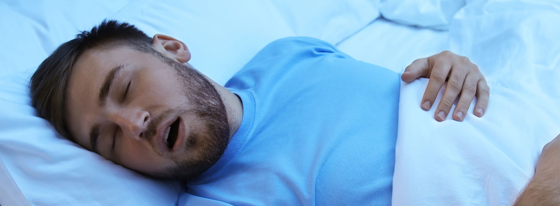 A man in a hospital bed, wearing a blue shirt and a white mask, appears to be asleep with his mouth open.