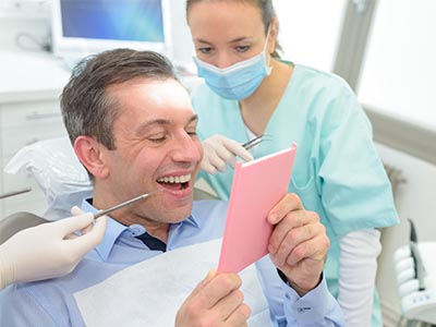 A man is sitting in a dental chair, holding up a pink card with a smile on his face while looking at it, surrounded by dental equipment and a smiling dentist.