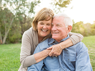 An elderly couple, the man is wearing a blue shirt and they are embracing each other in an outdoor setting.