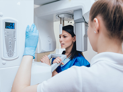 A medical professional in a blue uniform stands next to an advanced imaging machine with a woman wearing protective gloves, both in a clean, clinical setting.