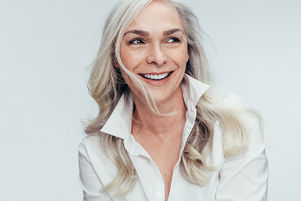 The image shows a woman with short hair smiling at the camera, wearing a white top and a silver necklace.