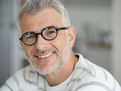 The image shows a man with gray hair, wearing glasses and a white shirt. He has a beard and mustache, and is smiling at the camera.