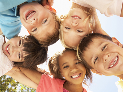 The image is a photograph of a group of children, likely taken outdoors during the daytime, with a clear blue sky in the background. They are smiling and appear to be enjoying themselves.
